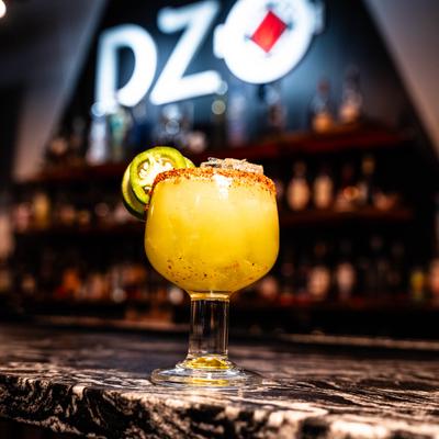 A spicy yellow margarita with jalapeño sits on a dark marble bar.