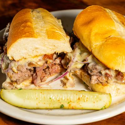 Tony’s Steak Sandwich served with a pickle spear on a white plate.