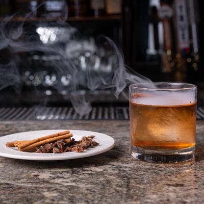 A smoking Old Fashioned sits on a bar top with a plate of star anise and cinnamon sticks.