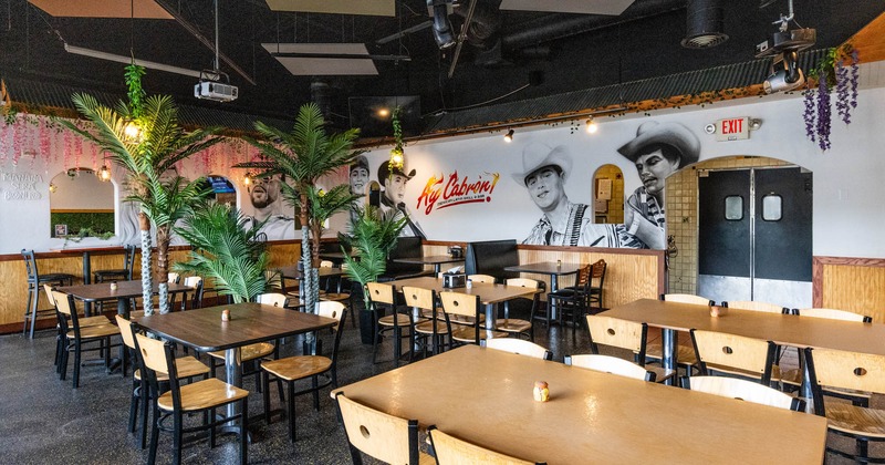 Interior of a restaurant with wooden tables, chairs, artificial palm trees, and wall murals
