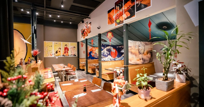 Interior of a sushi restaurant, with wooden tables, separate seating booths, plants and Japanese themed art on the walls
