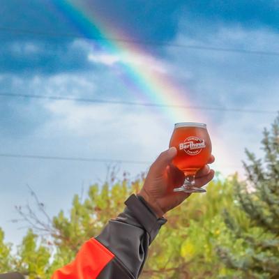 A hand raising a glass of red ale against a cloudy sky with a rainbow.