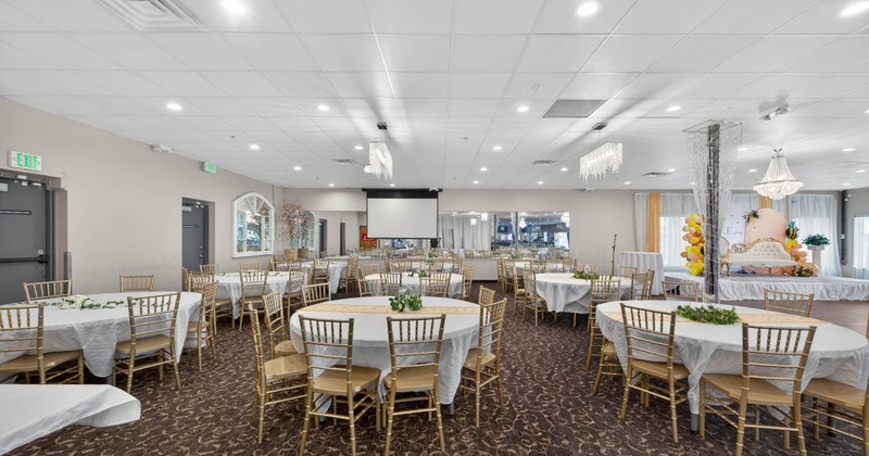 A large event hall with round tables, white tablecloths, gold chairs, chandeliers, and a stage with decorations