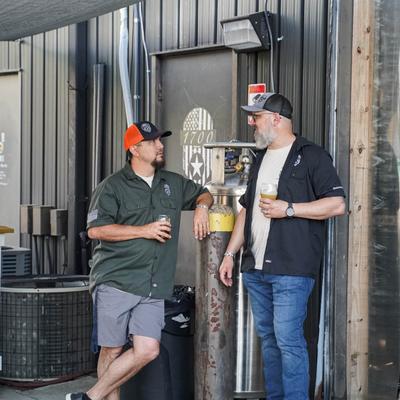 Two people are chatting outside a metal building, holding drinks, with relaxed expressions.