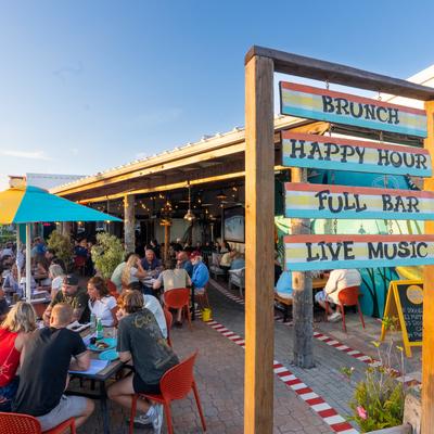 People dining on a restaurant patio beside a colorful sign.