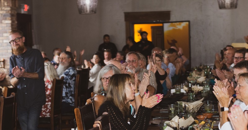 A group of people seated and standing at a long dining table, clapping and smiling