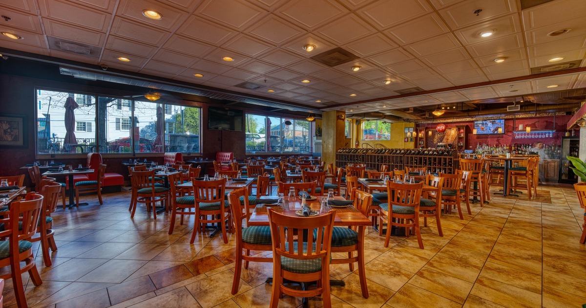 Spacious restaurant interior with wooden tables, chairs and a long bar in warm lighting.