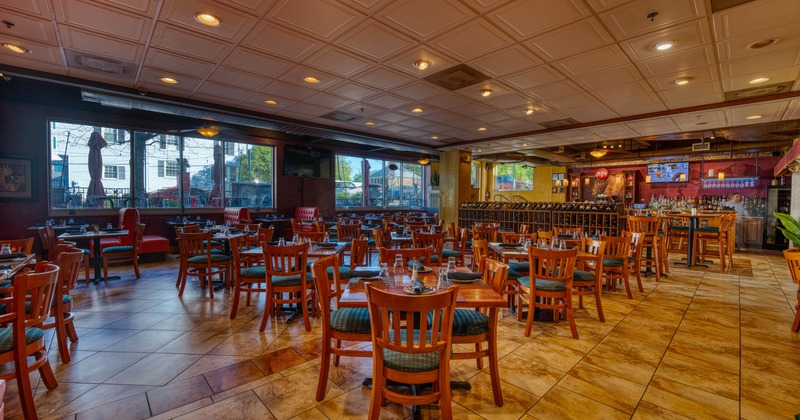 Spacious restaurant interior with wooden tables, chairs and a long bar in warm lighting.
