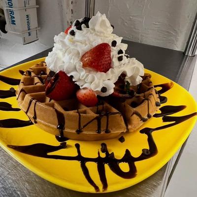 Waffle on a yellow plate, topped with strawberries, whipped cream, and chocolate drizzle.
