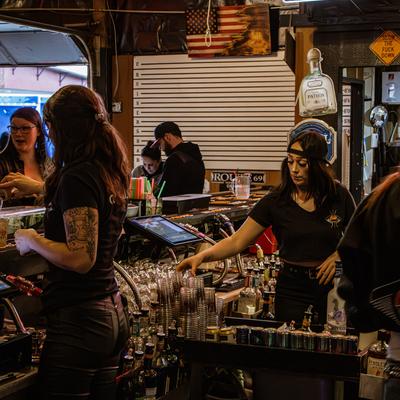 Area behind a bar, busy bartenders, guests, plastic cups, liquor bottles.