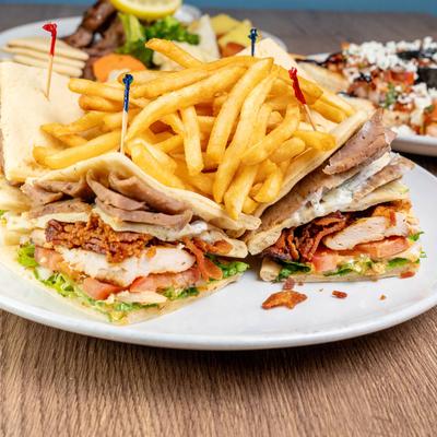 A white plate holds Athenian Club sandwich and fries on a wooden table.