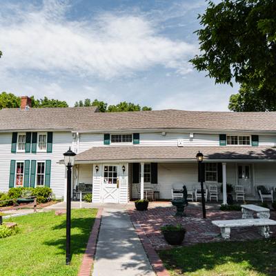 Exterior, white farmhouse style restaurant building with a lawn next to a paved walkway.