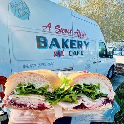 A freshly made turkey sandwich  is held up in front of the bakery's  truck.