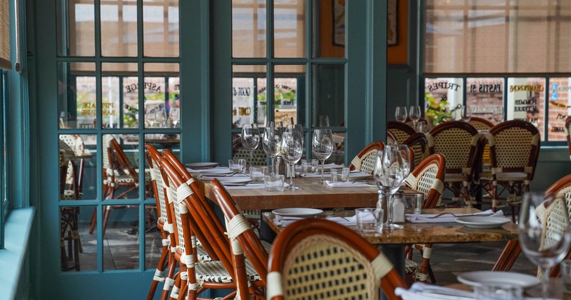 Restaurant dining room with teal glass doors, set tables, wine glasses, and woven cane chairs