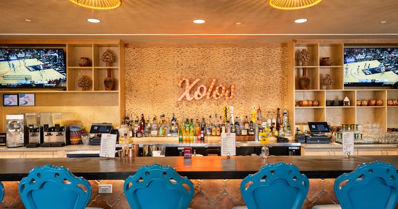 A bar with teal bar stools, alcohol bottles, TVs showing a basketball game and a neon sign Xolos