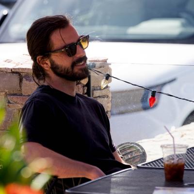 Person with a beard and sunglasses wearing a black t-shirt outdoors at a table with an iced drink.