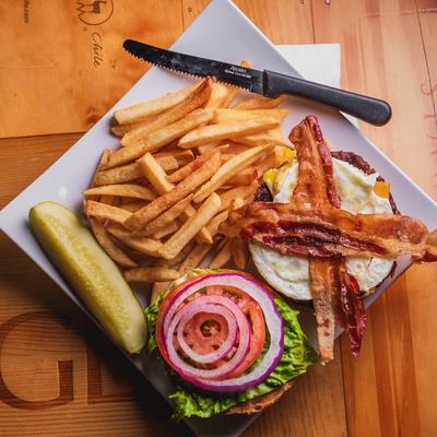 Burger with bacon, egg, lettuce, tomato, and red onion, served with fries and pickle.