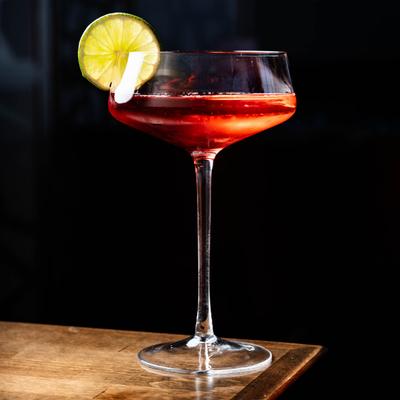 A red cocktail in a coupe glass, garnished with a lime slice.