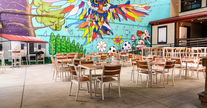 Outdoor seating area with wooden tables and chairs in front of a vibrant mural