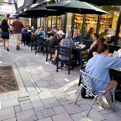 Patio dining area with people seated at tables under black umbrellas along a paved sidewalk.