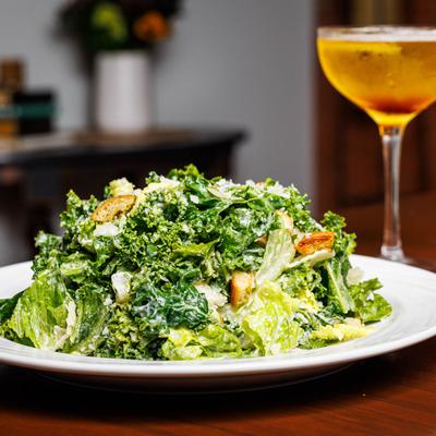 Caesar salad plate placed on a wooden table beside a tall glass of amber drink.
