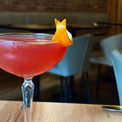 A red cocktail garnished with lemon peel sits on a table in dining area.