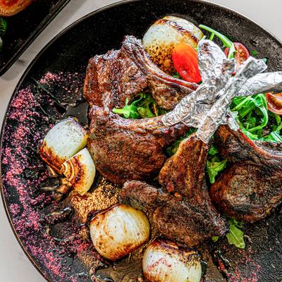 A plate holds grilled lamb chops, arugula salad with cherry tomatoes, and grilled onions.