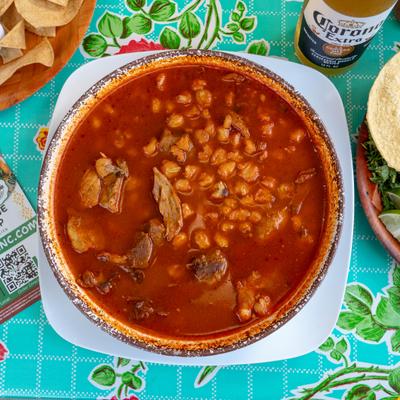 Bowl of Pozole Rojo served alongside fresh garnishes, a tortilla and a bottle of beer.