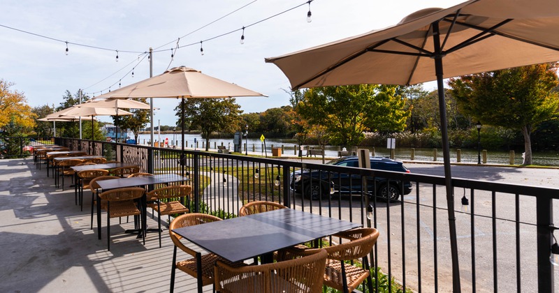 Outdoor dining area with tables, chairs, umbrellas, and string lights by a waterfront