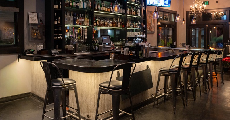A modern bar with a dark counter, high chairs, liquor shelves and hanging lights
