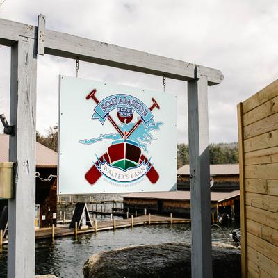 Sign for Squamside Beach Bar with logo, near wooden docks and lake.