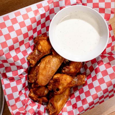Top-down view of chicken wings and a cup of ranch sauce served on checkered paper.