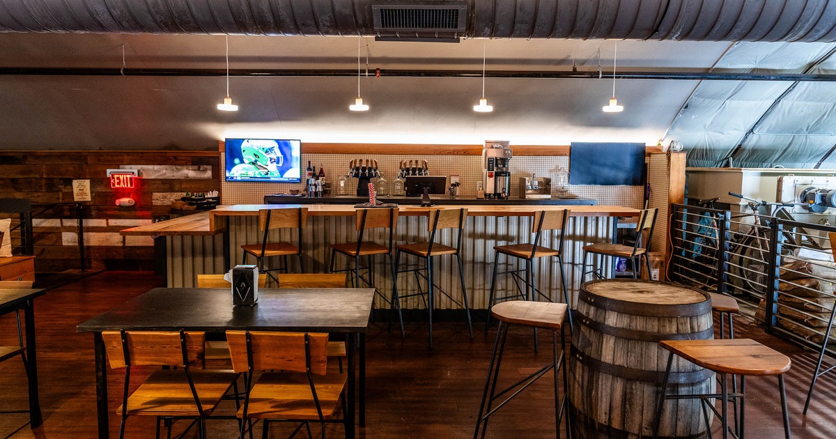 A rustic interior with a long counter, high stools, a TV and wooden tables and chairs.