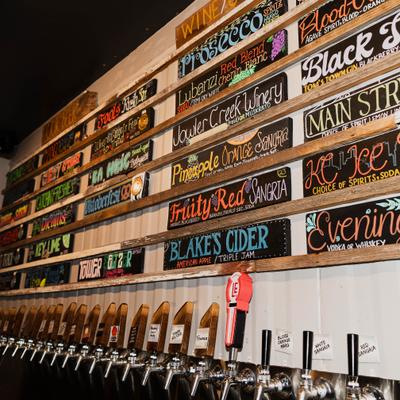 Wall of colorful chalkboard drink menus above a long row of beverage taps in a bar..
