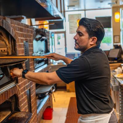 A cook uses a peel to place food into a brick oven in a bright kitchen.