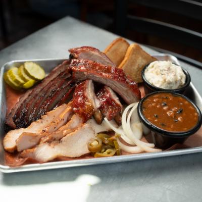 Barbecue platter with various meats and sides of baked beans, coleslaw and bread.
