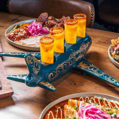 Set of four shots served in a decorative, airplane-shaped holder, alongside food.