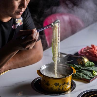 A person holds steaming noodles with chopsticks over a yellow hot pot on a table.