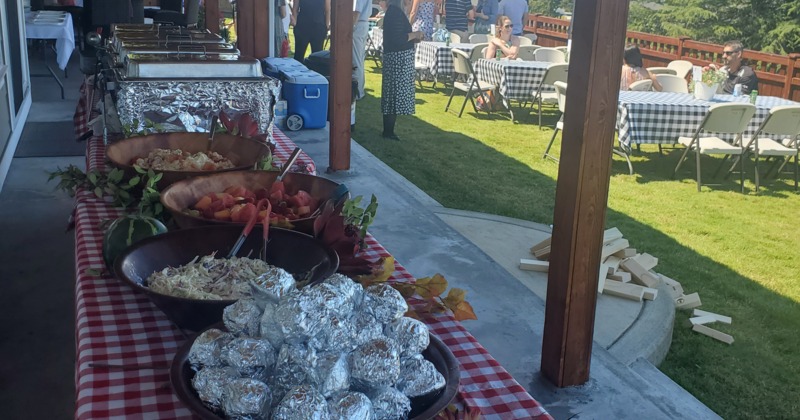 Outdoor buffet with foil-wrapped items, salads, and guests dining on lawn