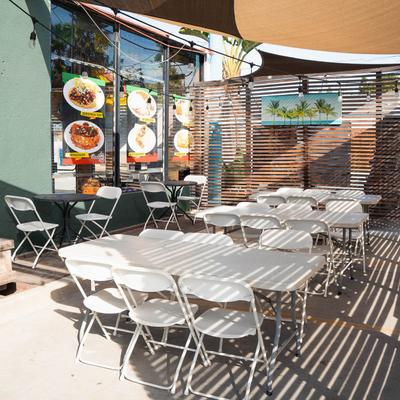 Outdoor dining area with white folding tables and chairs, shaded by a canopy.