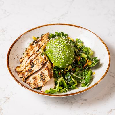A bowl contains grilled chicken and a variety of greens with sesame sprinkled avocado.