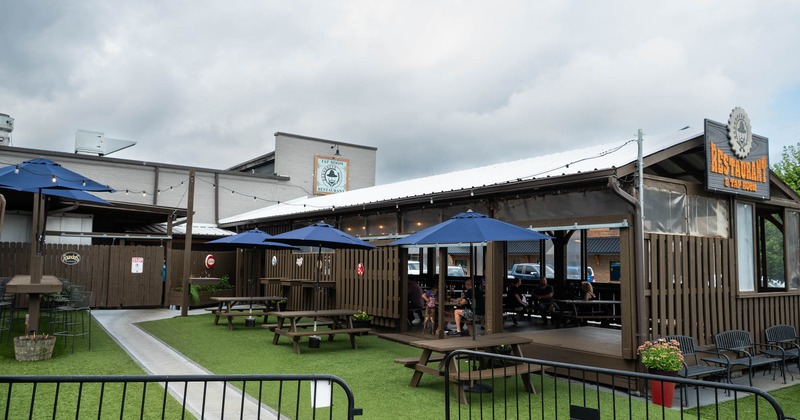 Outdoor seating area with picnic tables, blue umbrellas, and string lights under a cloudy sky