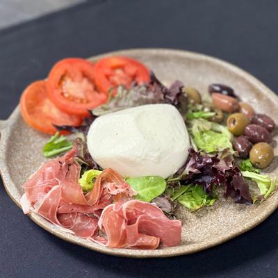 Burrata salad featuring tomatoes, prosciutto, mixed salad greens, and a side of olives.