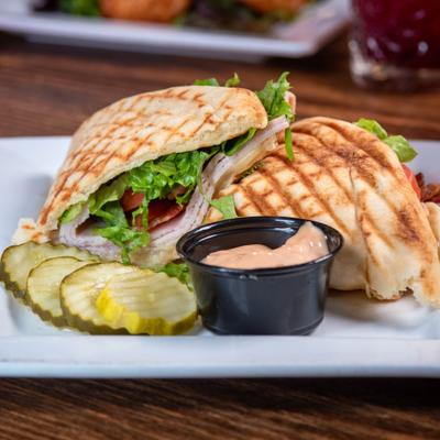 Turkey Club Panini served with pickle slices and a dipping sauce.