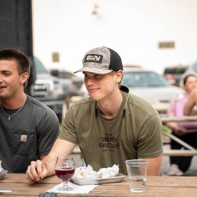 Young man sitting at an outdoor table with drinks and food trays, with people and cars in the background.