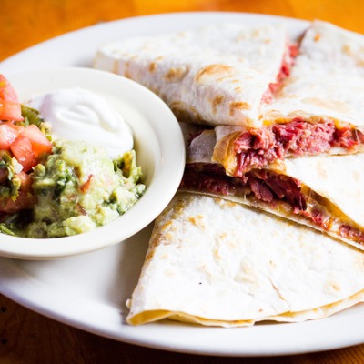 Quesadilla stuffed with corned beef on a plate served with a dish that contains guacamole, sour cream and pico de gallo