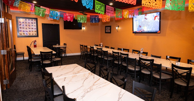 Dining area with long tables, chairs, colorful papel picado banners, and a TV on the wall