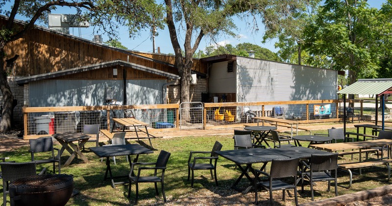Outdoor seating area with tables, chairs, and a grill in front of a shaded fence and buildings