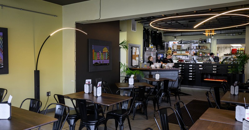 Inside, wooden tables, black chairs, a colorful city skyline artwork, and staff at a bar counter