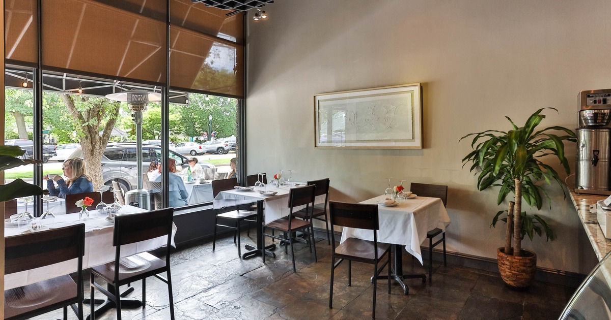 Interior of a restaurant with tables set with white tablecloths, near large windows showing outdoor seating and greenery